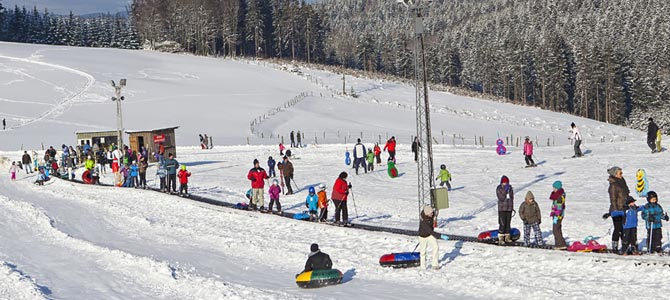 Auf einem verschneiten, von Bäumen umgebenen Hügel stehen Menschen Schlitten und warten in der Schlange, wobei ein kleines Gebäude und verschiedene bunte Schlitten zu sehen sind.
