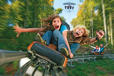 Zwei Kinder fahren auf einer Achterbahn in einem Waldgebiet, beide lächeln und schreien vor Aufregung; eine andere Gruppe ist im Hintergrund zu sehen.