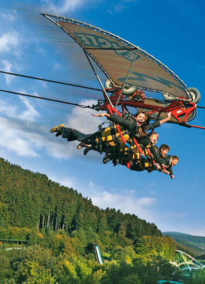 Sechs Personen schweben in einem Drachenflieger über einer bewaldeten Landschaft mit einem blauen Himmel im Hintergrund.