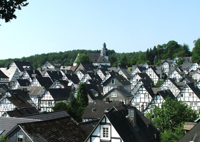 Ein Blick auf ein Dorf mit zahlreichen traditionellen Fachwerkhäusern und einer Kirche auf einem Hügel im Hintergrund, umgeben von Bäumen.