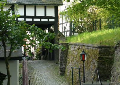 Ein kopfsteingepflasterter Weg führt unter einem Fachwerktor hindurch, das von moosbewachsenen Steinmauern und Grünpflanzen gesäumt ist. Rechts steht eine altmodische Lampe.