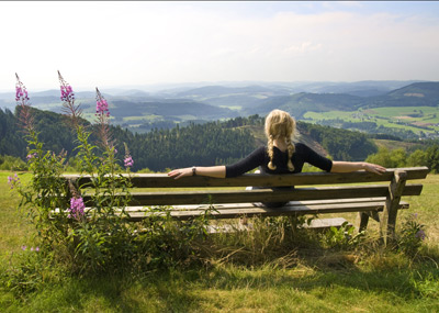 Eine Person mit blondem Haar sitzt auf einer Holzbank und blickt auf eine malerische Landschaft mit Hügeln, Wäldern und Feldern unter einem klaren Himmel.