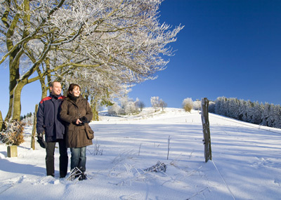 Ein Mann und eine Frau stehen zusammen auf einem schneebedeckten Feld unter blauem Himmel, mit Bäumen und einem Holzzaun in der Nähe.