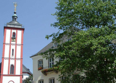 Ein rot-weiß gestreifter Kirchturm steht neben einem cremefarbenen Gebäude und einem großen, belaubten Baum unter einem klaren blauen Himmel.