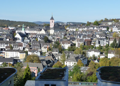 Ein Panoramablick auf eine europäische Stadt mit dichter Bebauung, einem markanten Kirchturm und den umliegenden grünen Hügeln unter einem klaren Himmel.
