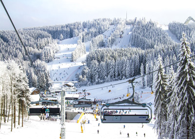 Skilift, der Menschen über ein verschneites Skigebiet befördert, mit Skifahrern auf den Pisten und schneebedeckten Bäumen im Hintergrund.