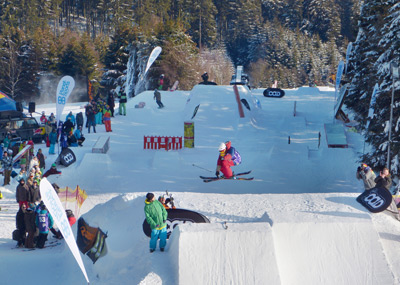 Ein Skifahrer, der in der Luft einen Sprung in einem verschneiten Geländepark ausführt, wobei im Hintergrund Zuschauer und Bäume zu sehen sind.