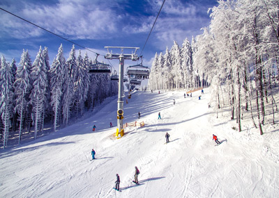 An einem klaren, sonnigen Wintertag fahren Skifahrer unter einem Skilift einen verschneiten Hang mit vereisten Bäumen hinunter.