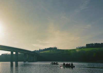 Mensen in kleine bootjes peddelen op een kalme rivier met een brug en groene heuvels op de achtergrond onder een heldere hemel.