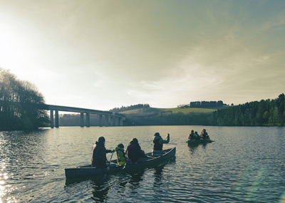 Drie kano's met mensen peddelen op een kalme rivier, met een brug en heuvels met bomen op de achtergrond onder een gedeeltelijk bewolkte hemel.