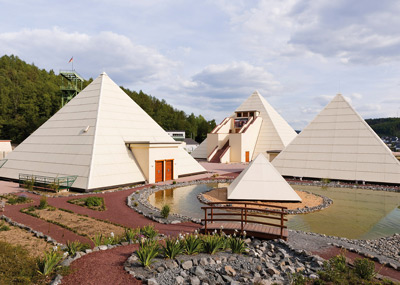 Several modern white pyramidal buildings are situated around a small pond with a wooden bridge, surrounded by trees and cloudy sky.