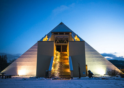 A large illuminated pyramid-shaped building with a central staircase covered in snow, photographed at dusk with a sign reading Einhorn above the entrance.
