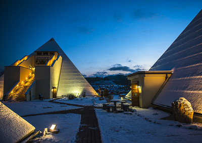 Snow-covered modern pyramid-shaped buildings at dusk, with illuminated stairs and a pathway leading between the structures.