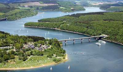 Luftaufnahme eines Flusses mit einer Brücke, Booten auf dem Wasser, grünen Wäldern, Feldern und Gebäuden entlang des Ufers.