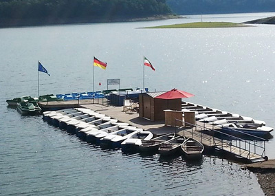 Een kleine aanlegsteiger met waterfietsen en roeiboten ligt voor anker in een kalm meer, met drie wapperende vlaggen en een klein schuurtje in het midden.