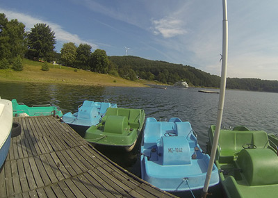 Verschillende groene en blauwe waterfietsen liggen aangemeerd aan een houten pier op een rustig meer, met een met gras begroeide, met bomen omzoomde oever en heuvels zichtbaar op de achtergrond.