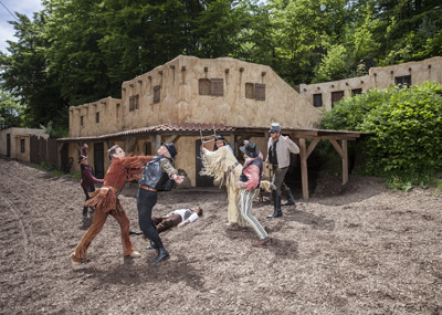 Een groep mensen gekleed in Wild West-kostuums doet alsof ze vechten bij een rustiek gebouw, waarbij één persoon op de grond ligt.