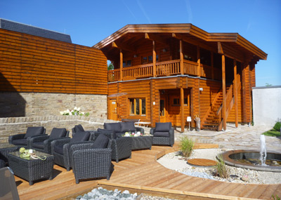 Een houten hut met twee verdiepingen, een balkon, rieten zithoek buiten, houten dek, kleine ronde vijver en heldere blauwe lucht.