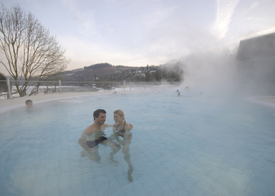 Een man en vrouw staan samen in een warmwaterbronbad omringd door stoom, met een winters landschap en kale bomen op de achtergrond.