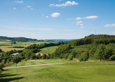 Een landelijk landschap met groene velden, verspreide bomen en heuvels in de verte onder een meestal helderblauwe lucht met een paar wolken.