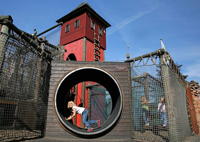 Een kind kruipt door een grote ronde tunnel op een buitenspeelplaats met een rode houten toren en touwbruggen op de achtergrond.