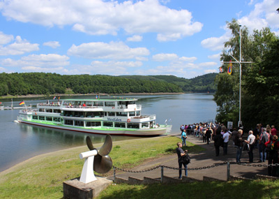 Een wit met groene passagiersboot ligt aangemeerd op een kalme rivier; een menigte mensen wacht aan wal onder een gedeeltelijk bewolkte hemel omringd door groen.