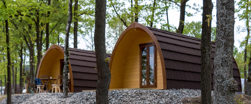 Zwei kleine, gewölbte Holzhütten mit braunen Dächern stehen zwischen Bäumen auf einem kiesbedeckten Gelände der Naturlodges, und in der Nähe einer Hütte sind Sitzgelegenheiten im Freien zu sehen.