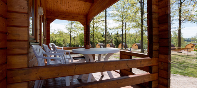 Houten veranda met witte plastic stoelen en een ronde tafel, met uitzicht op bomen en kleine houten Blockhaus-hutten op een camping.