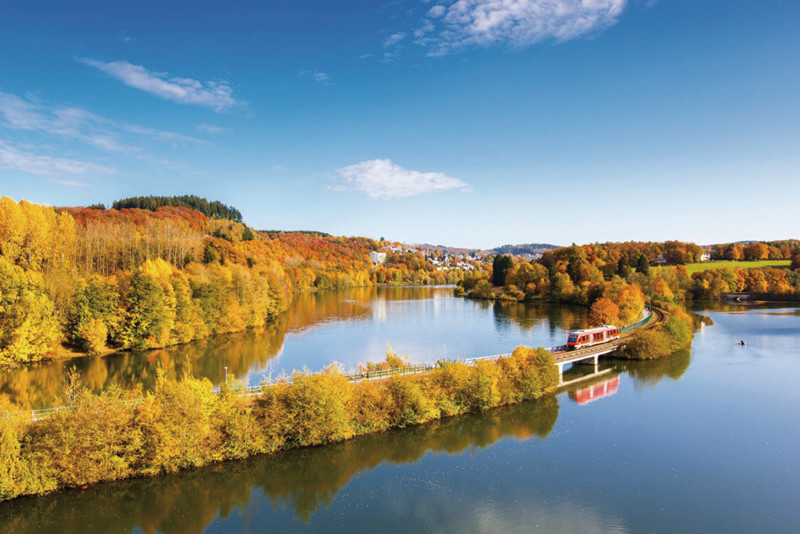 A train crosses a bridge over a calm river surrounded by trees with autumn foliage under a clear blue sky.