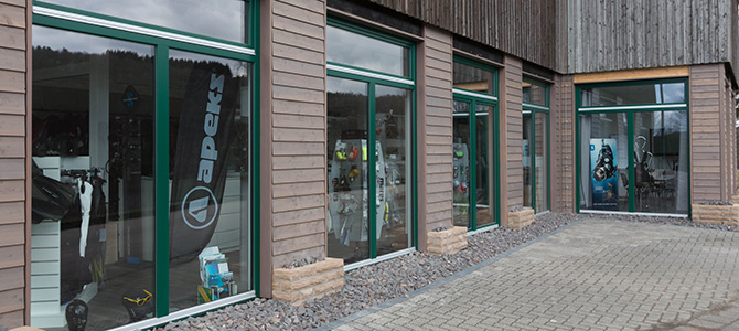Buitenaanzicht van een winkel met grote ramen met groene omlijsting die duikuitrusting tonen en reclamebanners binnenin.
