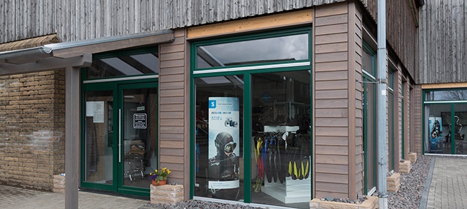 Buitenaanzicht van een moderne houten winkelpui met grote glazen etalages waarin outdoorkleding en -spullen worden uitgestald.