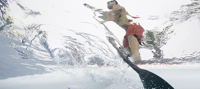 Unterwasseransicht einer Person, die auf einem Stand-up-Paddleboard paddelt, wobei das Wasser spritzt und das Paddel im Vordergrund zu sehen ist.