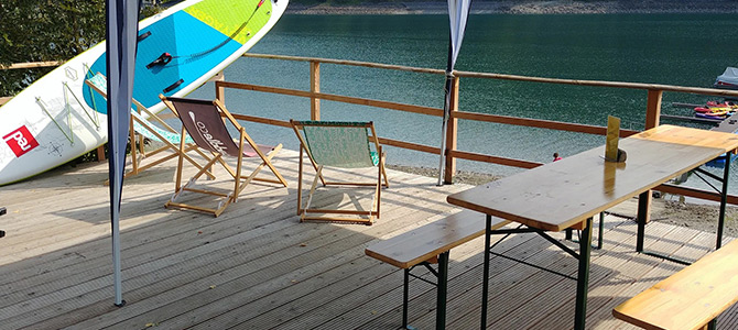 Eine Holzterrasse mit Klappstühlen, einem Picknicktisch und Bänken bietet einen Blick auf einen See; zwei Paddelbretter sind an das Geländer gelehnt.