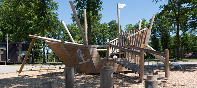 Een houten speelstructuur in de vorm van een schip met een helling, touwen en klimelementen, op mulch en omringd door bomen.