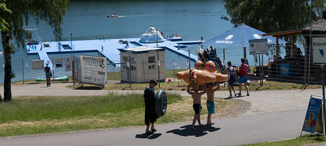 Mensen met springkussens lopen naar een waterpark aan het meer met drijvende constructies, terwijl anderen bij een kassa en een met paraplu's overdekt gedeelte staan.