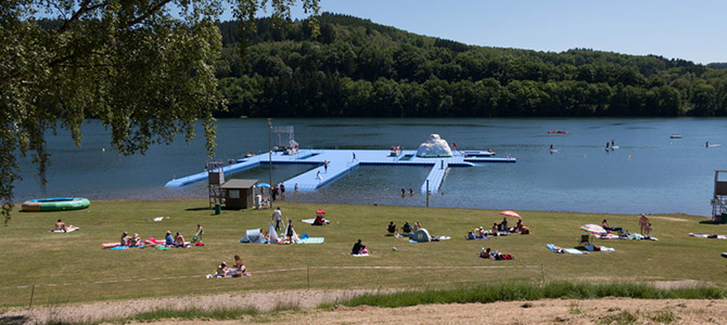 Mensen ontspannen op het gras bij een meer met een drijvend blauw platform en een waterglijbaan, omringd door bomen onder een heldere hemel.