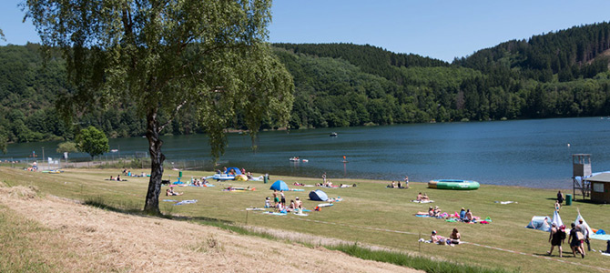 Mensen ontspannen op een zonnige dag op een grasstrand met handdoeken, parasols en springkussens, omringd door beboste heuvels en kalm water.