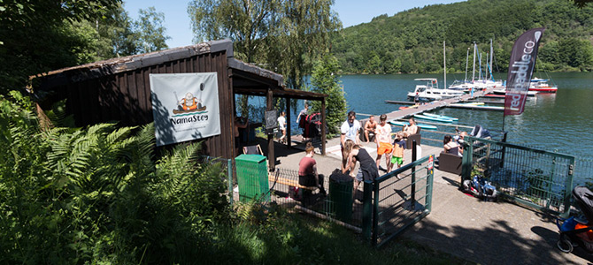 Eine Gruppe von Menschen versammelt sich vor einer Holzhütte in der Nähe einer Anlegestelle mit Booten auf einem See, umgeben von Bäumen und Hügeln. Ein Namasté9-Schild hängt an der Hütte.