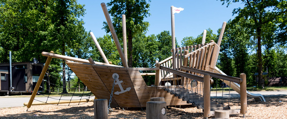 Spielplatz aus Holz in Form eines Schiffes mit Rutsche, Kletternetzen und Treppen. Dieser charmante Spielplatz ist von Bäumen umgeben und steht auf Holzspänen.
