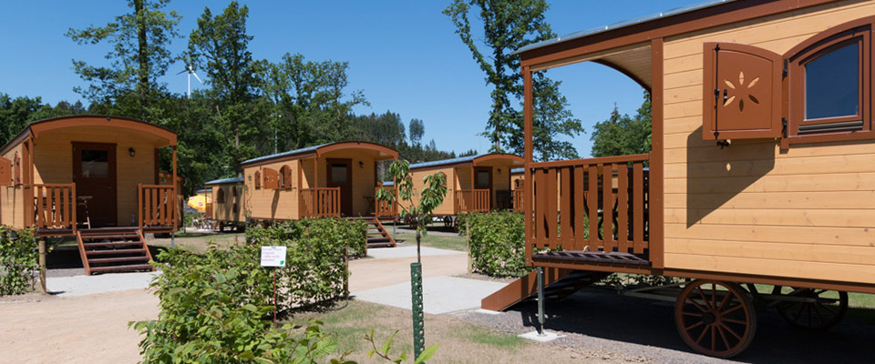 Mehrere kleine hölzerne Naturwagen mit Stufen und Vordächern stehen in einer Reihe auf einem Campingplatz, umgeben von Grün und Bäumen unter einem klaren blauen Himmel.