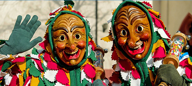 Twee mensen in kleurrijke kostuums met bladmotieven en houten maskers met overdreven gezichtsuitdrukkingen, de een zwaaiend, op wat een festival of parade lijkt te zijn.