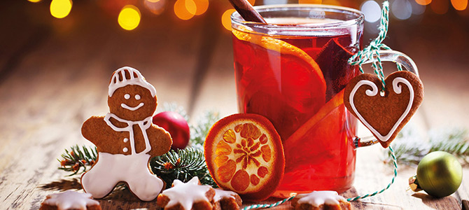 Een glazen mok glühwein met sinaasappelschijfjes en een kaneelstokje staat op een houten tafel, omringd door koekjes van peperkoek en kerstversieringen.