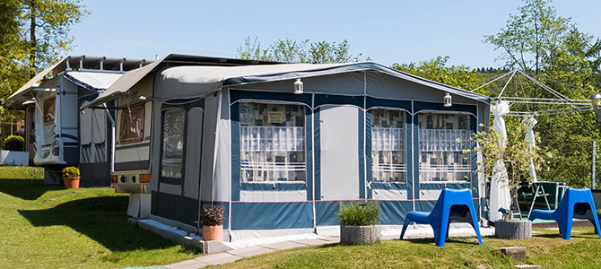 Een grote caravan met luifel staat op een grasveld, omringd door potplanten, blauwe stoelen en een klein terras onder een heldere, zonnige hemel.