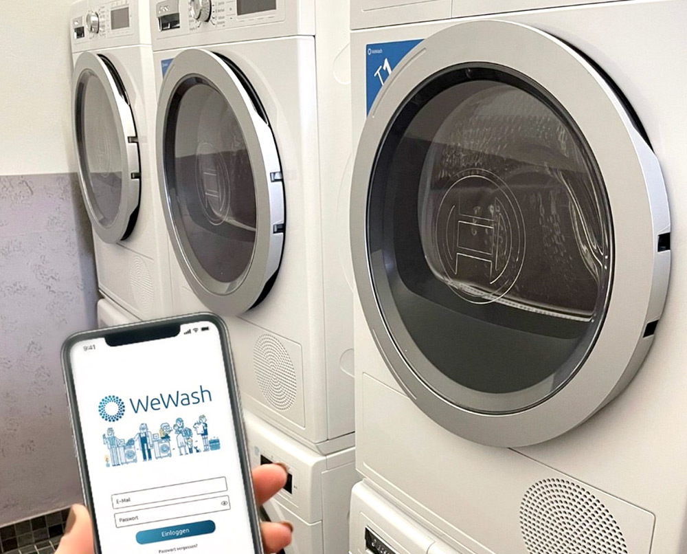 Eine Person hält ein Smartphone mit der WeWash-App vor eine Reihe von Frontlader-Waschmaschinen in einer Waschküche.