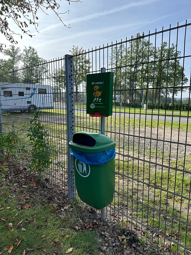 Ein grüner Hundekotbehälter mit einem blauen Müllbeutel und einem praktischen Spender für Abfallbeutel ist an einem Metallzaun in einem von Gras und Bäumen umgebenen Außenbereich angebracht.