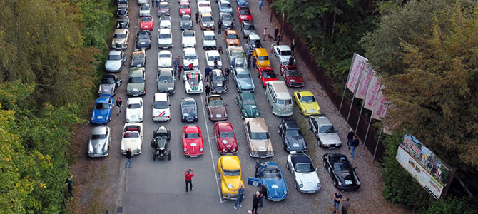 Luftaufnahme von mehreren Oldtimern, die in Reihen auf einer Straße geparkt sind, mit herumstehenden Menschen und Bäumen auf beiden Seiten.