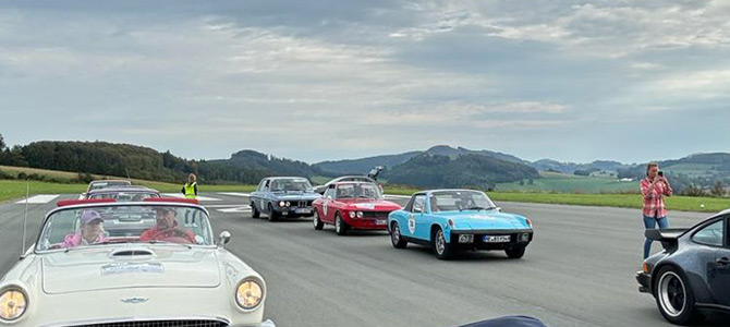 Eine Reihe von Oldtimern ist auf einer offenen Straße geparkt, Menschen stehen in der Nähe und im Hintergrund sind sanfte Hügel unter einem bewölkten Himmel zu sehen.