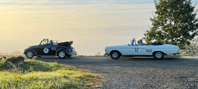 Zwei Oldtimer-Cabrios, ein schwarzes und ein weißes, sind während des Sonnenaufgangs oder Sonnenuntergangs auf einer gepflasterten Straße in der Nähe von Gras und Bäumen geparkt.