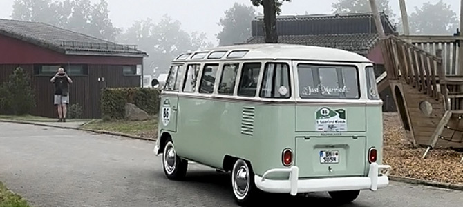 Ein hellgrün-weißer Oldtimer-Volkswagen fährt an einem nebligen Tag auf einer Straße in der Nähe von Häusern und einem Spielplatz.