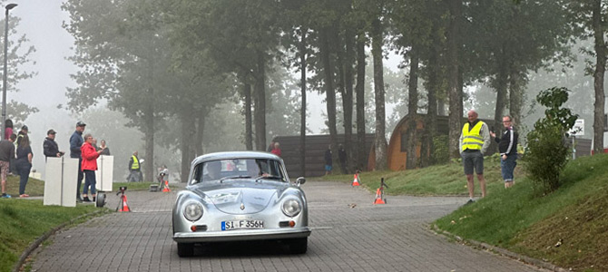 Ein silberner Oldtimer fährt während einer Veranstaltung im Freien auf einer asphaltierten Straße, an deren Rand Menschen in einer nebligen, baumbestandenen Gegend stehen.